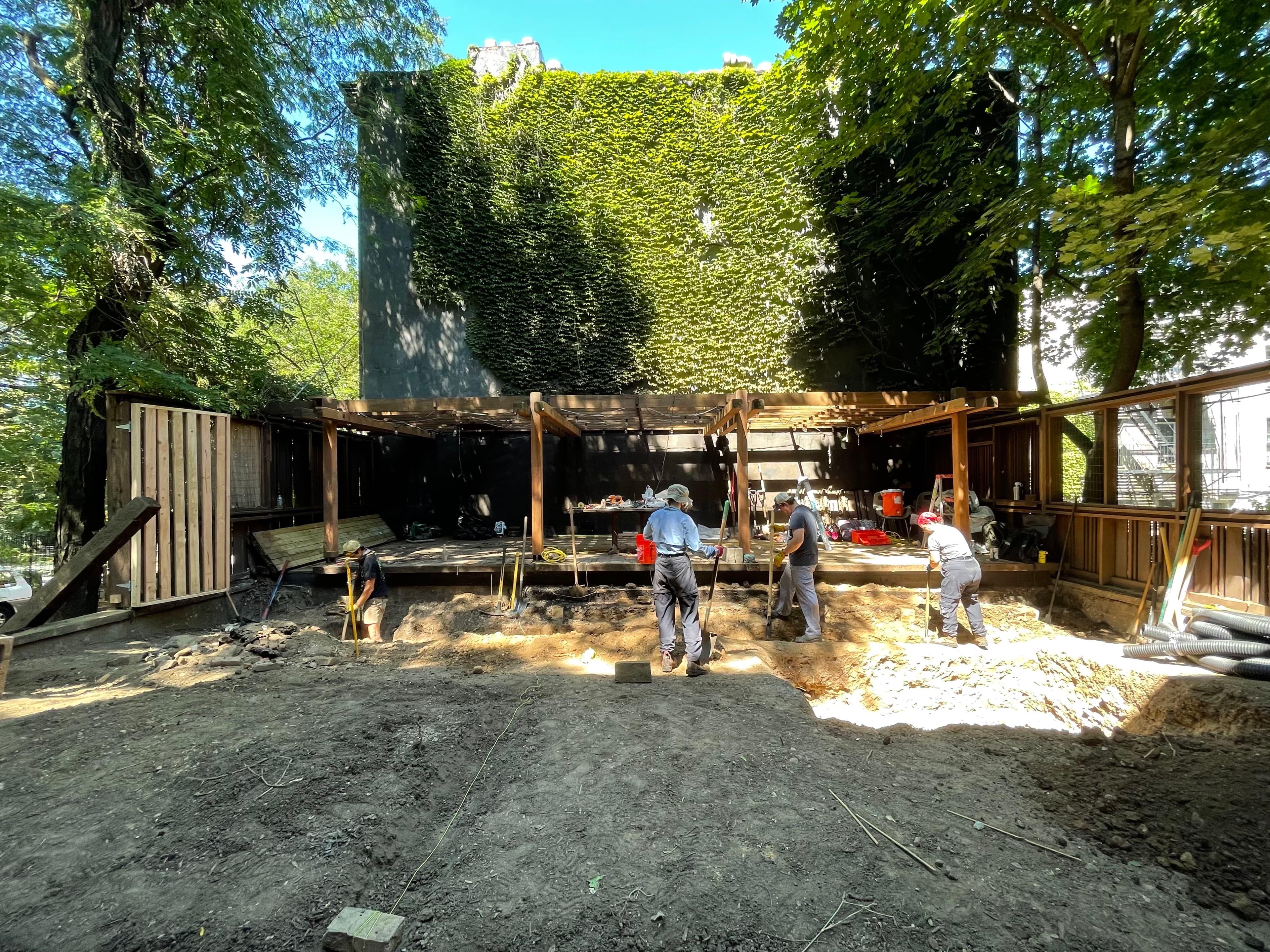 Installation in progress with a crew excavating the courtyard before the ivy-clad Merz House and its cedar pergola