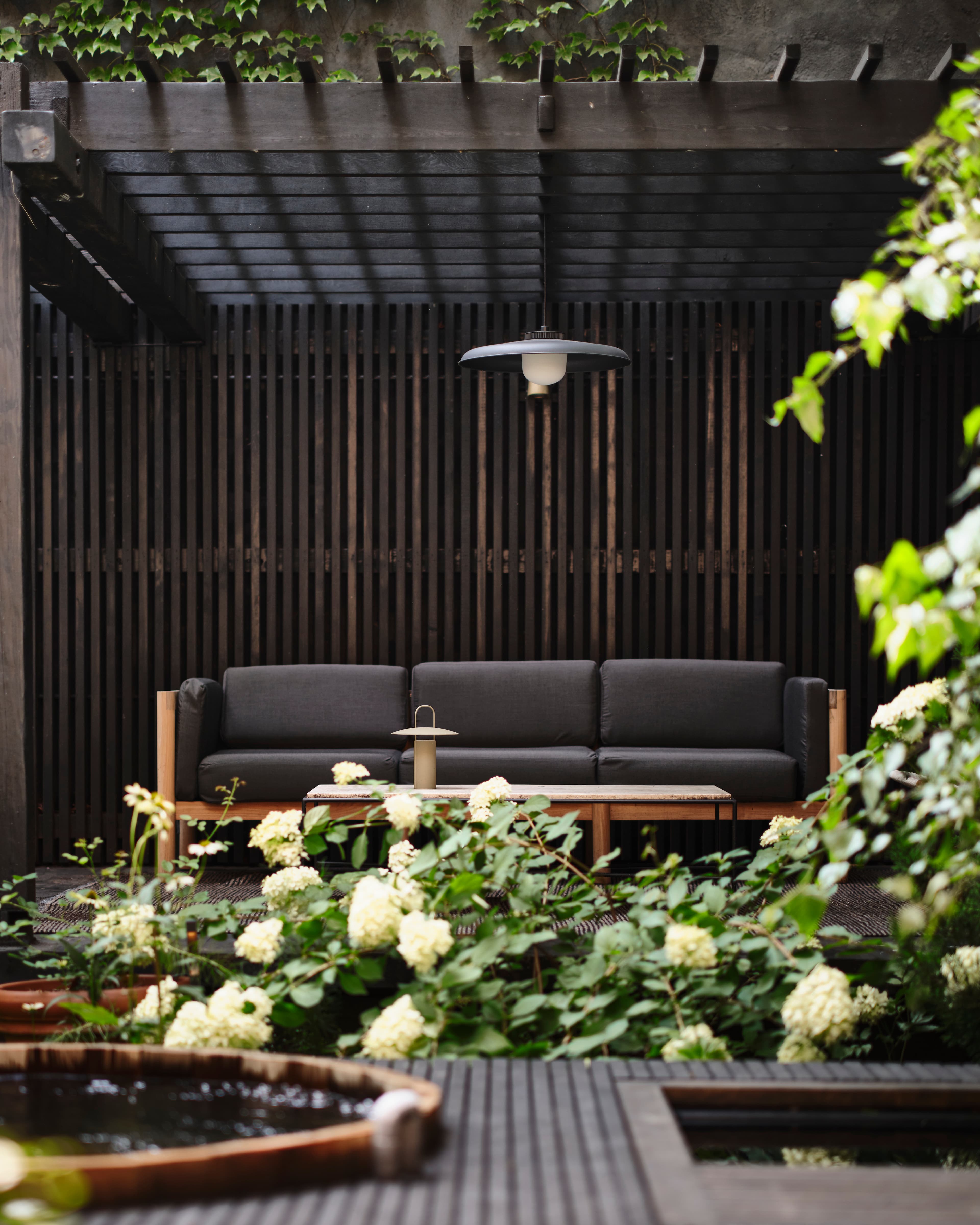 Shaded lounge beneath a blackened pergola with a dark sofa, framed by limelight hydrangea with the cedar hot tub in the foreground