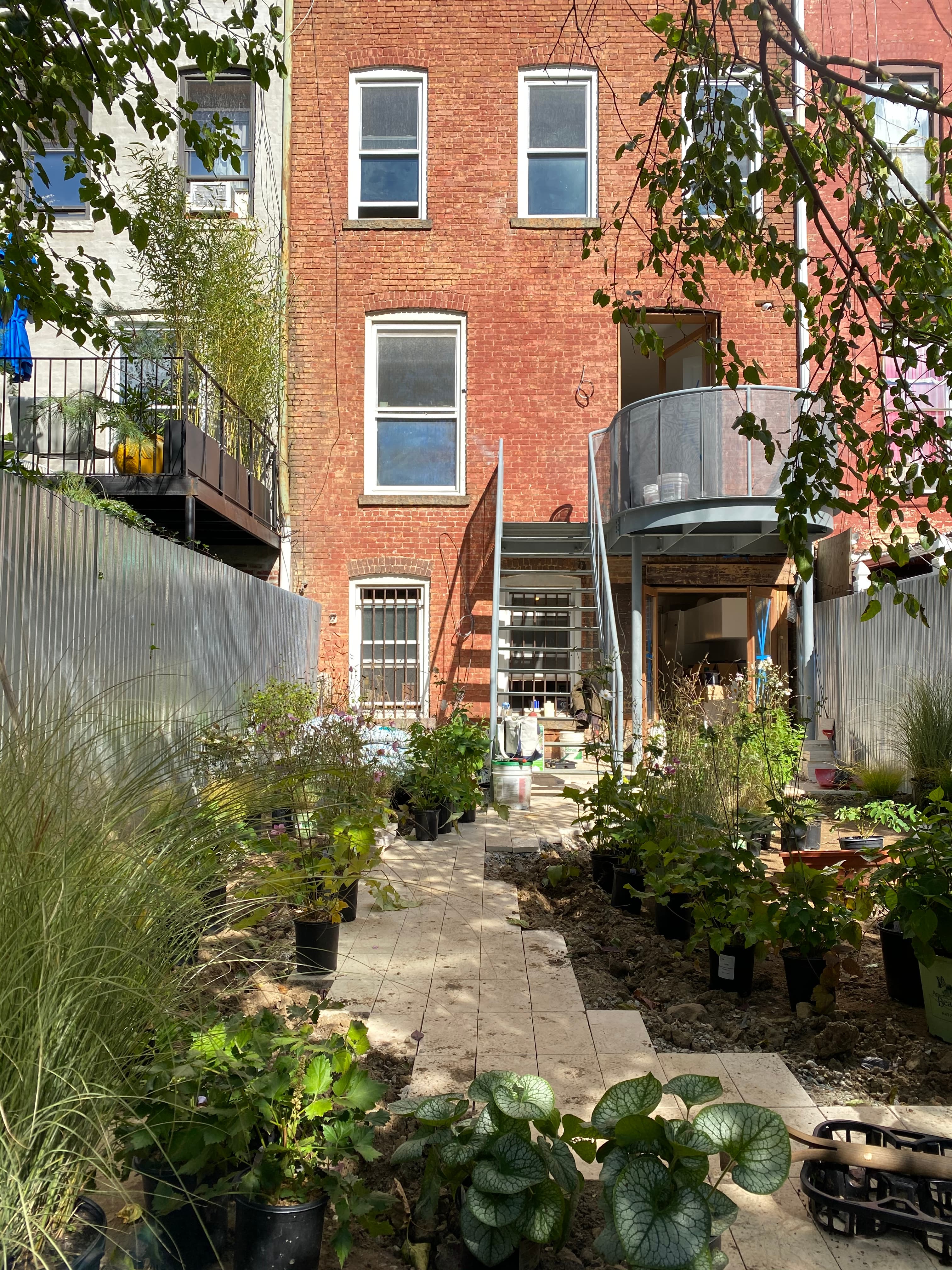 Installation in progress with a galvanized stair, freshly laid limestone paving, and staged nursery pots behind the brick brownstone